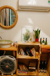 A cozy interior corner with a circular wicker-framed mirror reflecting a window with curtains. Below the mirror, a wooden shelf holds a variety of books and a framed botanical print. A potted plant on the shelf adds greenery. Above, an air conditioning unit is mounted on the wall. To the side, bottles of wine are neatly arranged, and a small fan sits near a sink with a decorative bowl.