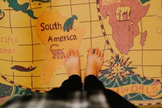 A textured world map rug featuring continents with an overhead view of a person's feet standing on South America. Animals are depicted on the continents, including an elephant on Africa and a monkey on South America. Earth-toned colors are prominently used.