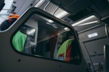 The interior of an aircraft is shown with a focus on the cockpit area. Two individuals are visible, both wearing fluorescent safety vests, likely indicating they are crew members or ground staff. Overhead, there are various panels and controls typical of an aircraft's interior. The lighting inside gives off a dim, bluish tone, creating a calm ambiance. An emergency exit sign is illuminated to the right.