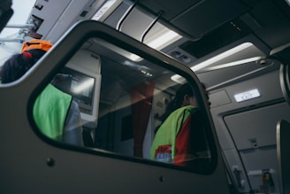 The interior of an aircraft is shown with a focus on the cockpit area. Two individuals are visible, both wearing fluorescent safety vests, likely indicating they are crew members or ground staff. Overhead, there are various panels and controls typical of an aircraft's interior. The lighting inside gives off a dim, bluish tone, creating a calm ambiance. An emergency exit sign is illuminated to the right.