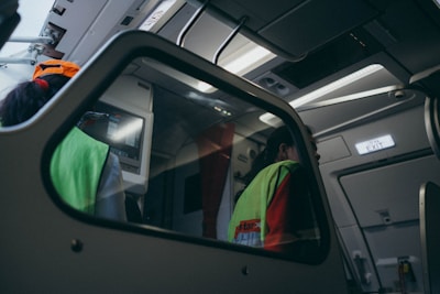 The interior of an aircraft is shown with a focus on the cockpit area. Two individuals are visible, both wearing fluorescent safety vests, likely indicating they are crew members or ground staff. Overhead, there are various panels and controls typical of an aircraft's interior. The lighting inside gives off a dim, bluish tone, creating a calm ambiance. An emergency exit sign is illuminated to the right.