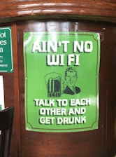 A poster on a wooden surface features a retro-style illustration of a man holding a beer mug. The poster is predominantly green with white text that reads 'AIN’T NO WI FI TALK TO EACH OTHER AND GET DRUNK.'