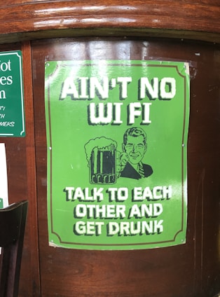 A poster on a wooden surface features a retro-style illustration of a man holding a beer mug. The poster is predominantly green with white text that reads 'AIN’T NO WI FI TALK TO EACH OTHER AND GET DRUNK.'
