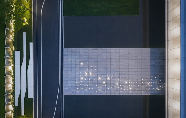 An aerial view captures a modern, geometric landscape design. A large, rectangular area is paved with tiles, illuminated by scattered lights that appear like stars on its surface. Adjacent to the tiles is a dark road with white lines, bordered by green grass and trimmed hedges. To the right, a building facade features vertical architectural elements, and to the left, landscaped greenery is outlined with more lighting.