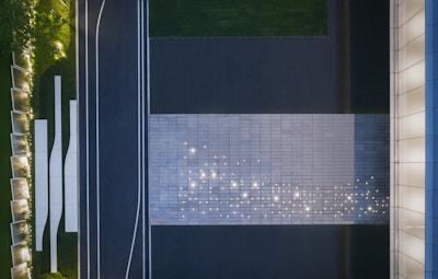 An aerial view captures a modern, geometric landscape design. A large, rectangular area is paved with tiles, illuminated by scattered lights that appear like stars on its surface. Adjacent to the tiles is a dark road with white lines, bordered by green grass and trimmed hedges. To the right, a building facade features vertical architectural elements, and to the left, landscaped greenery is outlined with more lighting.