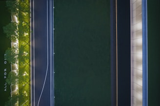 An aerial view of a landscaped area featuring a pathway flanked by trees with illuminated ground lighting. An adjacent road runs parallel, marked with white lines and dotted lights. A modern building with a sleek design is visible, showing a repetitive pattern of windows. The phrase 'ALL FOR ONE' is discernible on the ground.