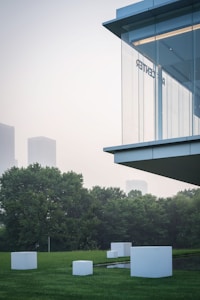 A modern building with glass walls is situated alongside a lawn with several white geometric sculptures. The background features lush green trees and the faint outline of tall city buildings, partially obscured by mist or fog.