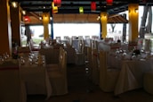 A dining space set up for an event, with tables arranged neatly and covered with white tablecloths. Each table is adorned with floral centerpieces and surrounded by chairs draped in white covers with pink sashes. Colorful lanterns hang from the ceiling, providing a festive ambiance.