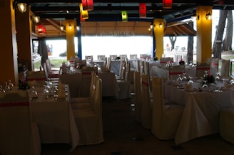 A dining space set up for an event, with tables arranged neatly and covered with white tablecloths. Each table is adorned with floral centerpieces and surrounded by chairs draped in white covers with pink sashes. Colorful lanterns hang from the ceiling, providing a festive ambiance.