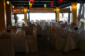 A dining space set up for an event, with tables arranged neatly and covered with white tablecloths. Each table is adorned with floral centerpieces and surrounded by chairs draped in white covers with pink sashes. Colorful lanterns hang from the ceiling, providing a festive ambiance.