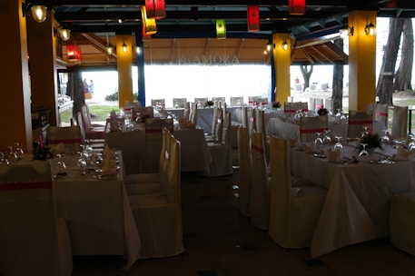 A dining space set up for an event, with tables arranged neatly and covered with white tablecloths. Each table is adorned with floral centerpieces and surrounded by chairs draped in white covers with pink sashes. Colorful lanterns hang from the ceiling, providing a festive ambiance.