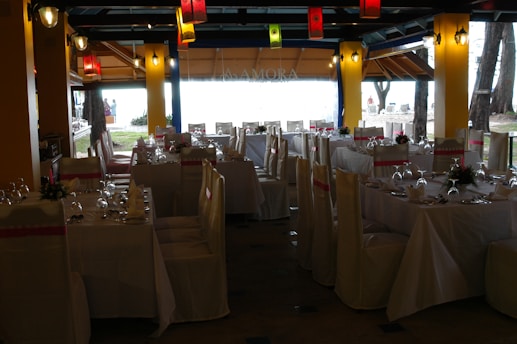 A dining space set up for an event, with tables arranged neatly and covered with white tablecloths. Each table is adorned with floral centerpieces and surrounded by chairs draped in white covers with pink sashes. Colorful lanterns hang from the ceiling, providing a festive ambiance.
