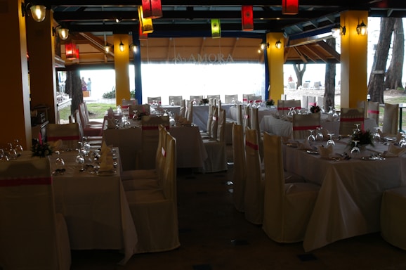 A dining space set up for an event, with tables arranged neatly and covered with white tablecloths. Each table is adorned with floral centerpieces and surrounded by chairs draped in white covers with pink sashes. Colorful lanterns hang from the ceiling, providing a festive ambiance.