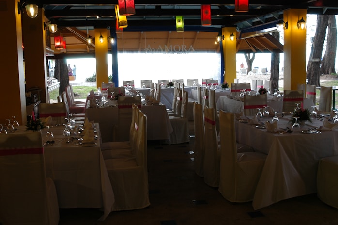 A dining space set up for an event, with tables arranged neatly and covered with white tablecloths. Each table is adorned with floral centerpieces and surrounded by chairs draped in white covers with pink sashes. Colorful lanterns hang from the ceiling, providing a festive ambiance.