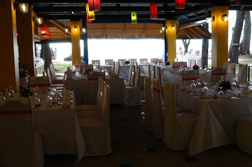 A dining space set up for an event, with tables arranged neatly and covered with white tablecloths. Each table is adorned with floral centerpieces and surrounded by chairs draped in white covers with pink sashes. Colorful lanterns hang from the ceiling, providing a festive ambiance.