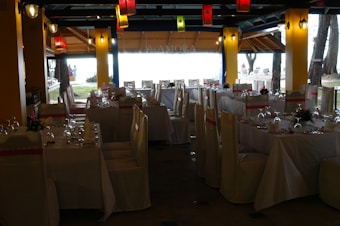 A dining space set up for an event, with tables arranged neatly and covered with white tablecloths. Each table is adorned with floral centerpieces and surrounded by chairs draped in white covers with pink sashes. Colorful lanterns hang from the ceiling, providing a festive ambiance.
