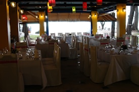 A dining space set up for an event, with tables arranged neatly and covered with white tablecloths. Each table is adorned with floral centerpieces and surrounded by chairs draped in white covers with pink sashes. Colorful lanterns hang from the ceiling, providing a festive ambiance.