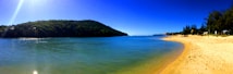 A pristine beach with golden sand curves gently alongside a calm, clear blue ocean under a vibrant blue sky. Dense, green foliage covers the hillside in the background. The sunlight casts a bright glow, adding a serene and inviting feel to the scene.