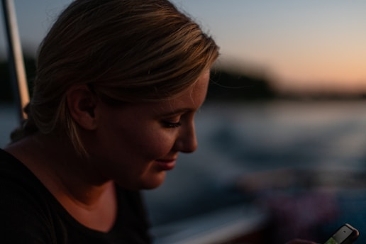 A woman with blonde hair is photographed in profile during sunset or twilight. She appears to be smiling slightly while looking at a smartphone in her hands. The blurred background suggests this might be taken on a body of water, with warm, soft lighting indicating an intimate atmosphere.
