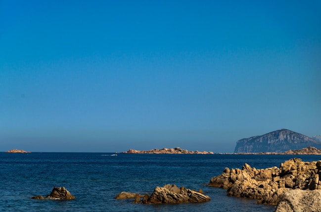 A serene ocean view with calm blue waters extending to the horizon. Rugged rocks frame the coastline, with distant landmasses and a prominent rocky hill visible against a clear blue sky.