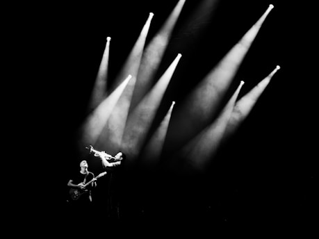 A musician playing a trumpet is illuminated under several bright stage lights, creating dramatic beams against a dark background. Another musician with a guitar stands nearby, contributing to the performance.