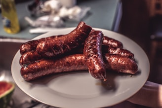 A plate filled with several grilled sausages, arranged neatly on a kitchen counter. The sausages are cooked to a golden brown, with a glossy sheen suggesting they are freshly prepared. A hint of blurry kitchen background including a bottle and some kitchen appliances is visible, indicating a casual home setting.
