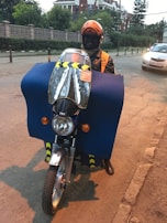 A person wearing an orange helmet and jacket is riding a motorcycle. The motorcycle is covered with a large blue protective cloth, and there is a reflective windshield attached to the front. A car is visible on the street, and buildings with green foliage are in the background.