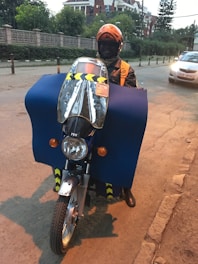 A person wearing an orange helmet and jacket is riding a motorcycle. The motorcycle is covered with a large blue protective cloth, and there is a reflective windshield attached to the front. A car is visible on the street, and buildings with green foliage are in the background.