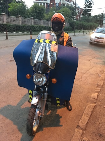 A person wearing an orange helmet and jacket is riding a motorcycle. The motorcycle is covered with a large blue protective cloth, and there is a reflective windshield attached to the front. A car is visible on the street, and buildings with green foliage are in the background.