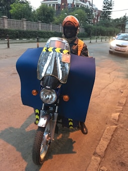 A person wearing an orange helmet and jacket is riding a motorcycle. The motorcycle is covered with a large blue protective cloth, and there is a reflective windshield attached to the front. A car is visible on the street, and buildings with green foliage are in the background.