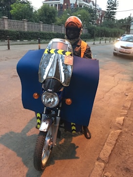 A person wearing an orange helmet and jacket is riding a motorcycle. The motorcycle is covered with a large blue protective cloth, and there is a reflective windshield attached to the front. A car is visible on the street, and buildings with green foliage are in the background.