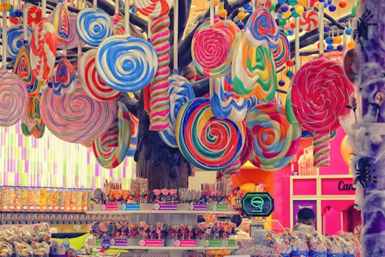 Giant colorful lollipops are suspended from the ceiling, displaying a variety of swirling patterns in vibrant hues. The background includes a candy store with shelves filled with various sweets, and two people are seen browsing the selection.