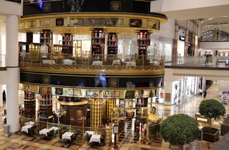 A luxurious, multi-level tea store and cafe with ornate metalwork and glass walls located in a shopping mall. The store interior is filled with tea canisters in various colors, and there are tables set up outside for dining. The surrounding area includes other retail stores with bright lighting and polished floors.