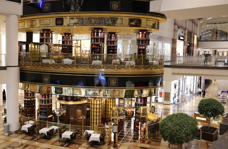 A luxurious, multi-level tea store and cafe with ornate metalwork and glass walls located in a shopping mall. The store interior is filled with tea canisters in various colors, and there are tables set up outside for dining. The surrounding area includes other retail stores with bright lighting and polished floors.