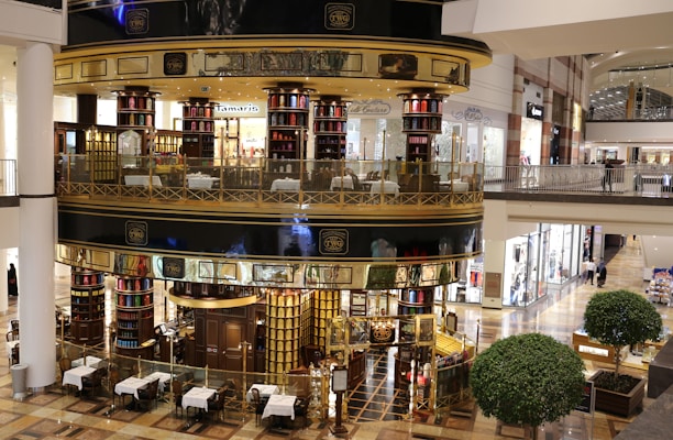 A luxurious, multi-level tea store and cafe with ornate metalwork and glass walls located in a shopping mall. The store interior is filled with tea canisters in various colors, and there are tables set up outside for dining. The surrounding area includes other retail stores with bright lighting and polished floors.