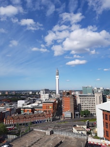 A cityscape with a prominent telecommunications tower rising above a mix of modern and older brick buildings. The sky is blue with scattered fluffy clouds, and the city appears bustling yet orderly. Roads and parking facilities are visible in the foreground.