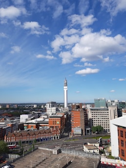 A cityscape with a prominent telecommunications tower rising above a mix of modern and older brick buildings. The sky is blue with scattered fluffy clouds, and the city appears bustling yet orderly. Roads and parking facilities are visible in the foreground.