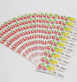 A collection of labels arranged in a fanned-out pattern, each featuring illustrations of a red factory with a green chimney and a yellow area with text and barcodes. The labels are neatly organized on a white background.