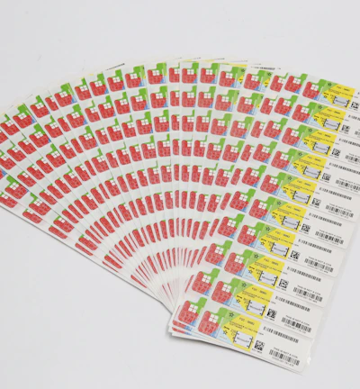 A collection of labels arranged in a fanned-out pattern, each featuring illustrations of a red factory with a green chimney and a yellow area with text and barcodes. The labels are neatly organized on a white background.