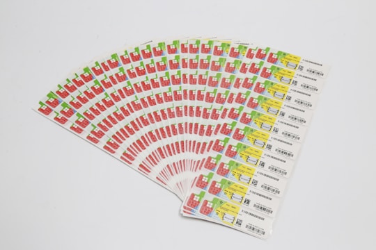 A collection of labels arranged in a fanned-out pattern, each featuring illustrations of a red factory with a green chimney and a yellow area with text and barcodes. The labels are neatly organized on a white background.