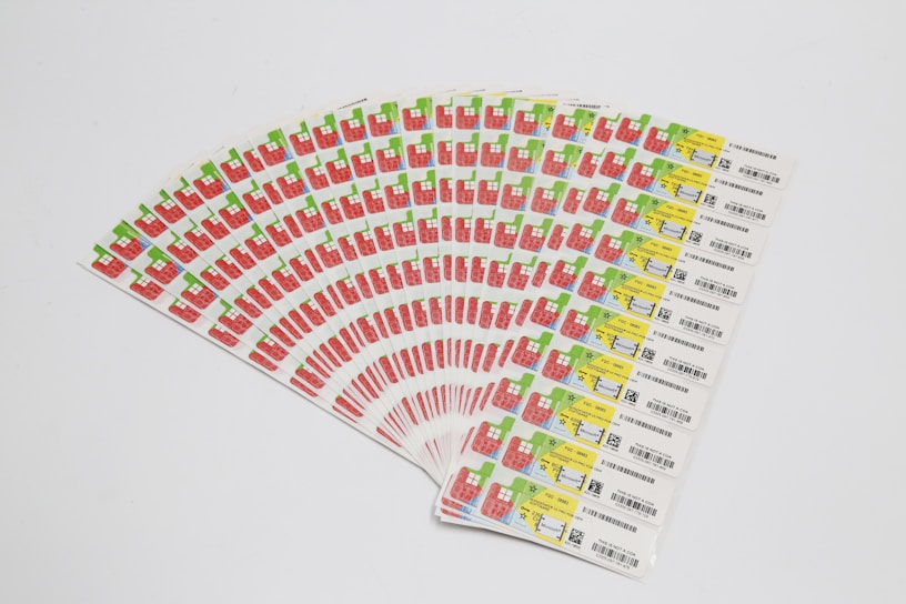 A collection of labels arranged in a fanned-out pattern, each featuring illustrations of a red factory with a green chimney and a yellow area with text and barcodes. The labels are neatly organized on a white background.