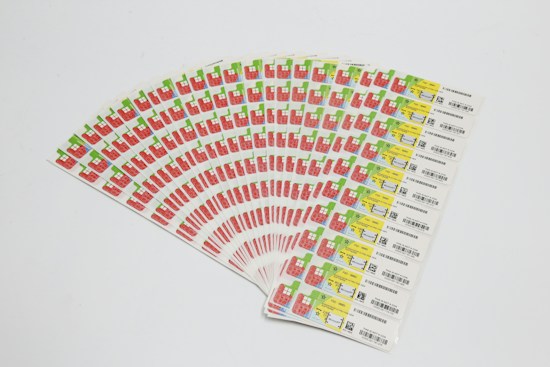 A collection of labels arranged in a fanned-out pattern, each featuring illustrations of a red factory with a green chimney and a yellow area with text and barcodes. The labels are neatly organized on a white background.