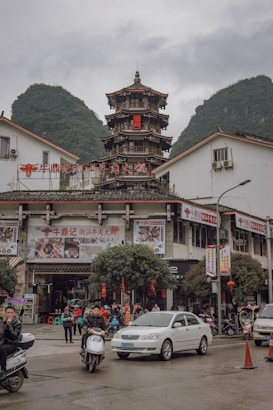 A busy street scene in front of a traditional multi-tiered Chinese tower, surrounded by modern white buildings with red signage. People are riding scooters and driving cars. The setting is framed by green, mist-covered mountains under a cloudy sky.