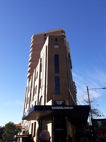 A tall building with a modern architectural design stands against a bright blue sky. The structure features clean lines and a prominent facade with windows. The signage indicates there are function rooms available, and there is a website displayed at the bottom.