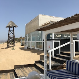 A beachside structure with glass walls and a thatched roof watchtower next to sand dunes. The area features wooden steps and seating with blue and white striped cushions, surrounded by clear blue skies.