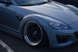 A close-up view of the front side of a sleek, modern car with a smooth, light blue exterior. The image highlights the large, metallic rimmed wheels and detailed headlights.