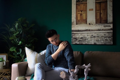 A person wearing a blue sweater and glasses is sitting on a beige sofa, gently resting their hand on their shoulder. The setting includes a dark green wall with a photograph of a street scene featuring two wooden doors and cobblestones. There are lush green plants on a side table and a cozy ambiance is created by the decorative elements.