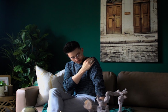 A person wearing a blue sweater and glasses is sitting on a beige sofa, gently resting their hand on their shoulder. The setting includes a dark green wall with a photograph of a street scene featuring two wooden doors and cobblestones. There are lush green plants on a side table and a cozy ambiance is created by the decorative elements.