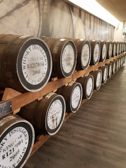 Rows of wooden whisky barrels are lined up on racks along a corridor. Each barrel has labels indicating 'Whisky Distillery King Car, Yuan Shan' with production numbers and dates. The barrels are placed in a well-lit and orderly fashion, suggesting a controlled environment for aging spirits.