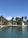 A luxurious waterfront house surrounded by tropical palm trees, with a clear blue sky overhead. The house is situated next to a calm waterway, with a small dock extending into the water. A white sailboat is moored nearby, adding to the serene and affluent ambiance.